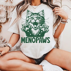 May include: Cream-colored t-shirt featuring a green graphic of a cat with its mouth open and paws outstretched. The word "MENOPAWS" is printed in green below the cat illustration. The shirt has short sleeves.