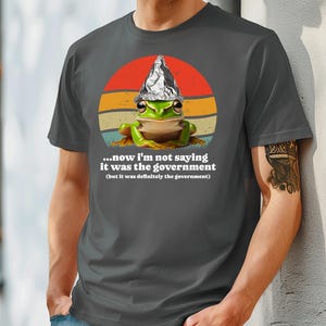 May include: Dark grey t-shirt with a cartoon frog wearing a foil hat, set against a retro sunset design. The text reads, "...now I'm not saying it was the government (but it was definitely the government)."