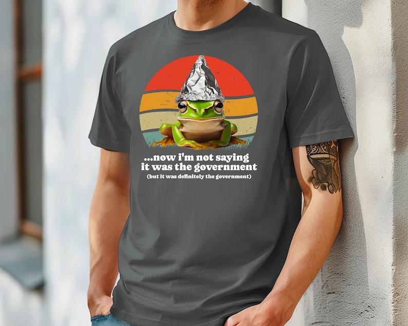 May include: Dark grey t-shirt with a cartoon frog wearing a foil hat, set against a retro sunset design. The text reads, "...now I'm not saying it was the government (but it was definitely the government)."