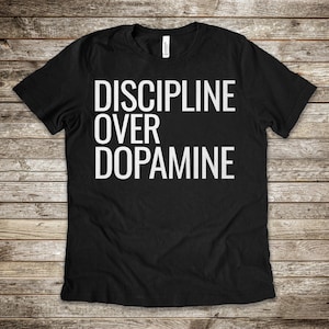 Puede incluir: Camiseta negra con el texto blanco "DISCIPLINE OVER DOPAMINE" en negrita, con fuente sans-serif. La camiseta se muestra sobre una superficie de madera.