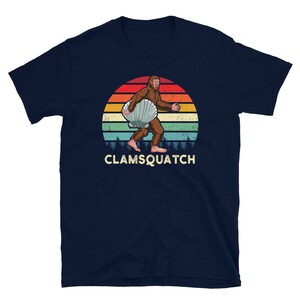 May include: A navy blue t-shirt with a retro-style illustration of a Bigfoot holding a seashell. The illustration is set against a sunset background with a vintage color palette. The text "CLAMSQUATCH" is printed below the illustration.