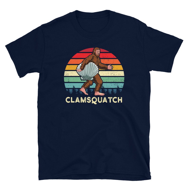 May include: A navy blue t-shirt with a retro-style illustration of a Bigfoot holding a seashell. The illustration is set against a sunset background with a vintage color palette. The text "CLAMSQUATCH" is printed below the illustration.