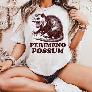 May include: Cream-colored t-shirt featuring a detailed illustration of an opossum with its mouth open. The text "PERIMENO POSSUM" is printed in a dark brown color below the opossum graphic. The shirt is a casual, unisex style.