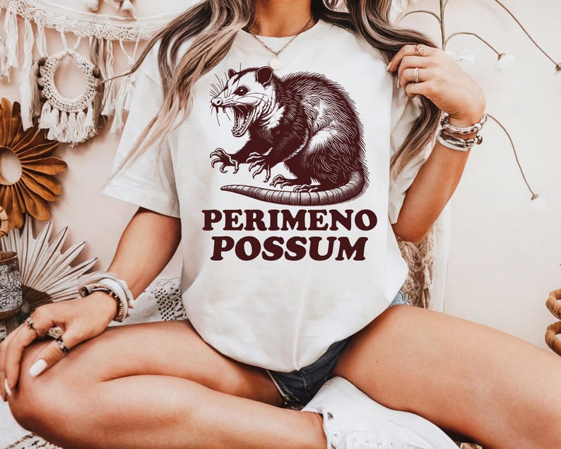 May include: Cream-colored t-shirt featuring a detailed illustration of an opossum with its mouth open. The text "PERIMENO POSSUM" is printed in a dark brown color below the opossum graphic. The shirt is a casual, unisex style.