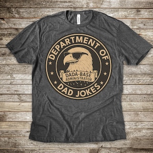 May include: Dark gray t-shirt with a circular graphic featuring an eagle wearing sunglasses. The graphic reads "DEPARTMENT OF DAD JOKES" around the edge, with "DADA-BASE ADMINISTRATOR" in the center. The shirt is made of a soft material.