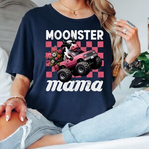May include: Navy blue t-shirt with the text "Moonster Mama". The graphic features a cow driving a pink monster truck with flowers in the back. The background is a pink and white checkered pattern.