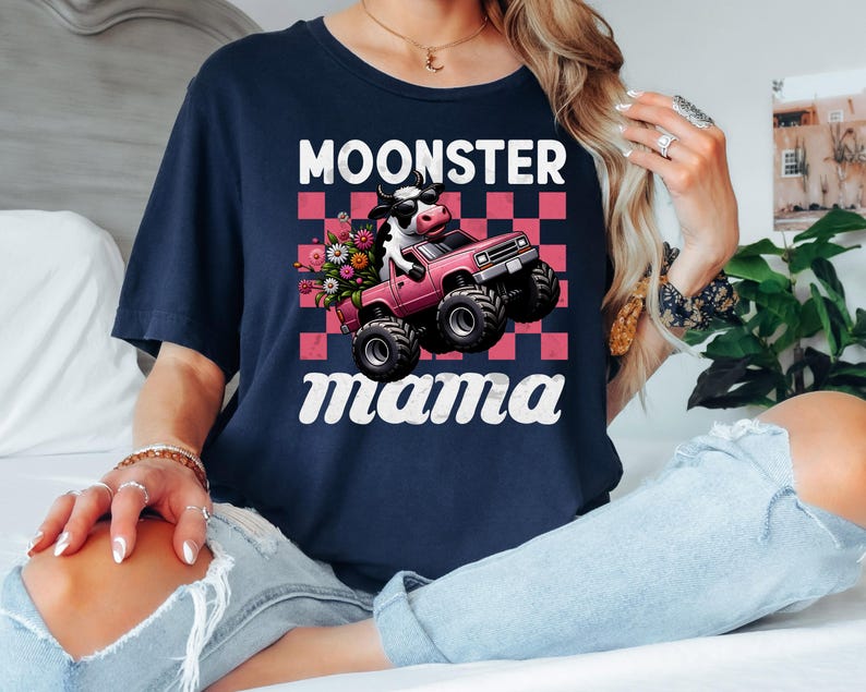 May include: Navy blue t-shirt with the text "Moonster Mama". The graphic features a cow driving a pink monster truck with flowers in the back. The background is a pink and white checkered pattern.