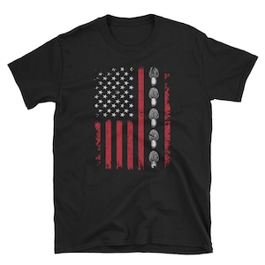 以下が含まれることがあります： 赤、白、青のアメリカ国旗のデザインが施された黒いTシャツ。旗は灰色のキノコの列で縁取られています。