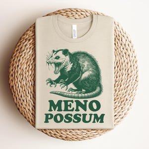 Pu&ograve; includere: Una maglietta beige con un'illustrazione verde di un opossum con la bocca aperta. Il testo "MENO POSSUM" &egrave; stampato sotto. La maglietta &egrave; piegata su un sottobicchiere rotondo e intrecciato.