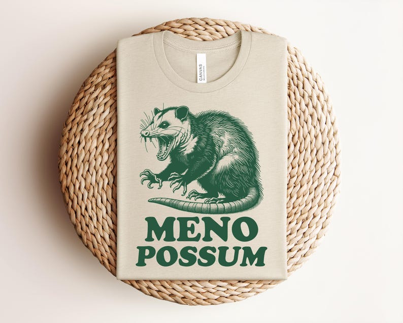 Pu&ograve; includere: Una maglietta beige con un'illustrazione verde di un opossum con la bocca aperta. Il testo "MENO POSSUM" &egrave; stampato sotto. La maglietta &egrave; piegata su un sottobicchiere rotondo e intrecciato.