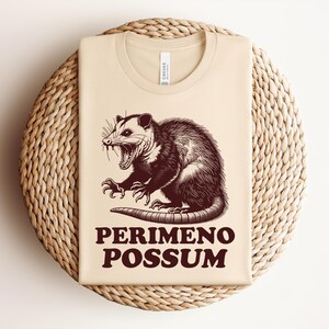 May include: Beige t-shirt with a brown illustration of an opossum with its mouth open and claws extended. The text "PERIMENO POSSUM" is printed in brown below the opossum. The shirt is folded on a woven, circular surface.