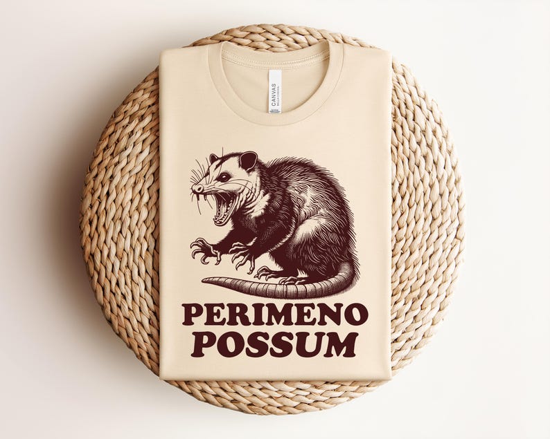 May include: Beige t-shirt with a brown illustration of an opossum with its mouth open and claws extended. The text "PERIMENO POSSUM" is printed in brown below the opossum. The shirt is folded on a woven, circular surface.