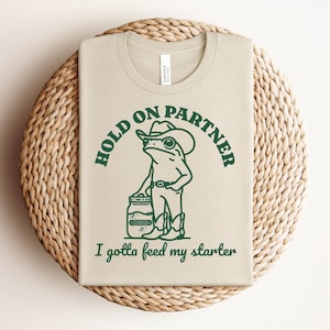 May include: Beige t-shirt with a green graphic of a frog wearing a cowboy hat and boots, holding a jar. Text reads "HOLD ON PARTNER" and "I gotta feed my starter."