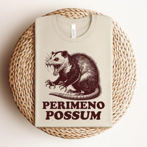 May include: Beige t-shirt with a brown illustration of an opossum with its mouth open and claws extended. The text "PERIMENO POSSUM" is printed in brown below the opossum. The shirt is folded on a woven, circular placemat.