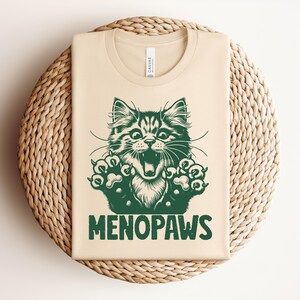 May include: A beige t-shirt with a green graphic of a cat with its mouth open and paws extended. The text "MENOPAWS" is printed below the cat illustration. The shirt is folded on a woven, circular surface.