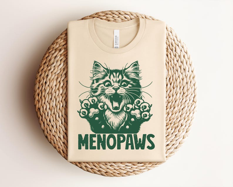 May include: A beige t-shirt with a green graphic of a cat with its mouth open and paws extended. The text "MENOPAWS" is printed below the cat illustration. The shirt is folded on a woven, circular surface.