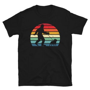 K&ouml;nnte beinhalten: Schwarzes T-Shirt mit einer Retro-Grafik eines Bigfoots, der einen Hund vor einem Sonnenuntergang spazieren f&uuml;hrt.