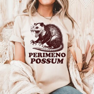May include: Beige t-shirt featuring a detailed illustration of an opossum with its mouth open, and the text "PERIMENO POSSUM" in a bold, dark brown font. The opossum graphic is intricately shaded, giving it a vintage, hand-drawn appearance.