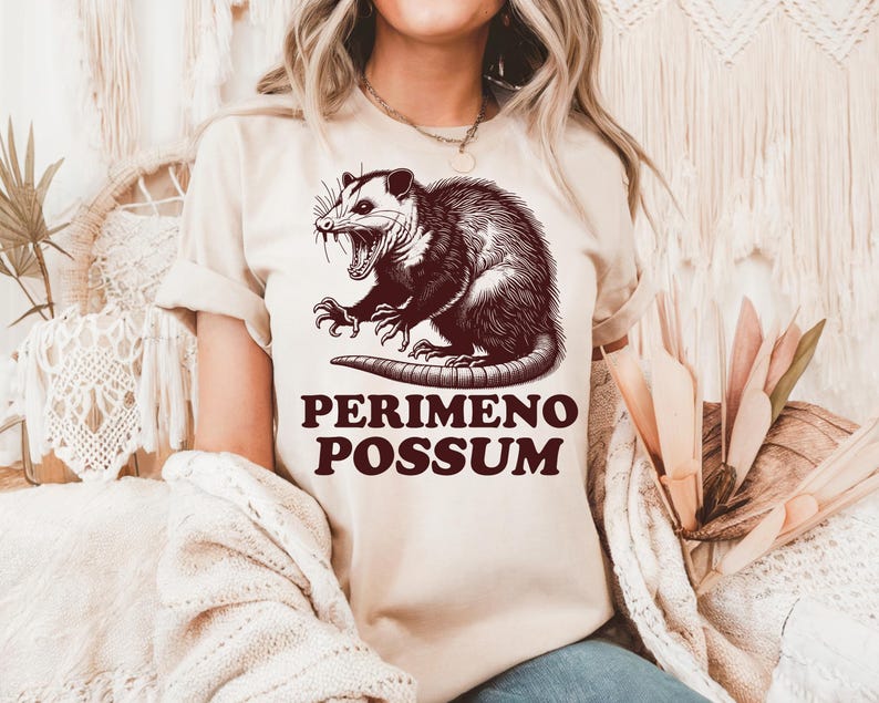 May include: Beige t-shirt featuring a detailed illustration of an opossum with its mouth open, and the text "PERIMENO POSSUM" in a bold, dark brown font. The opossum graphic is intricately shaded, giving it a vintage, hand-drawn appearance.