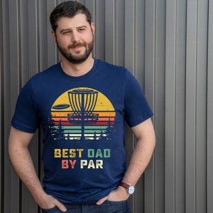 May include: A navy blue t-shirt with a vintage-style graphic of a disc golf basket and the text "Best Dad By Par".