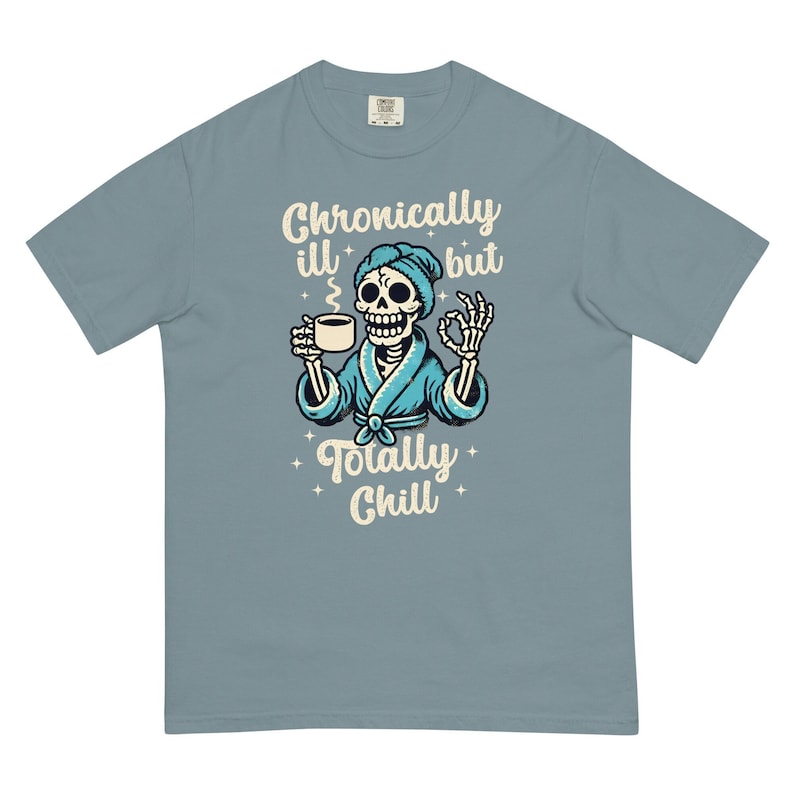 K&ouml;nnte beinhalten: Ein hellblaues T-Shirt mit einem Cartoon-Skelett, das einen blauen Bademantel tr&auml;gt und eine Tasse Kaffee h&auml;lt. Der Text auf dem Shirt lautet "Chronically ill but totally chill".