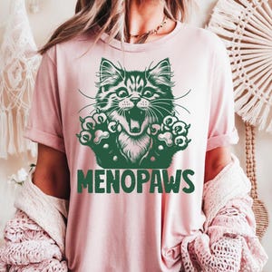 May include: A light pink t-shirt featuring a green illustration of a cat with its mouth open and paws extended. The text "MENOPAWS" is printed in green below the cat graphic. The shirt has short sleeves.