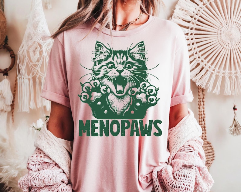 May include: A light pink t-shirt featuring a green illustration of a cat with its mouth open and paws extended. The text "MENOPAWS" is printed in green below the cat graphic. The shirt has short sleeves.