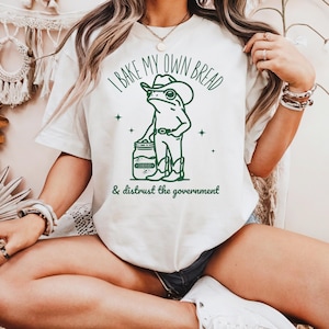 Puede incluir: Camiseta color crema con un gráfico verde de una rana con sombrero y botas de vaquero, sosteniendo un tarro. El texto dice "I BAKE MY OWN BREAD" y "& distrust the government".