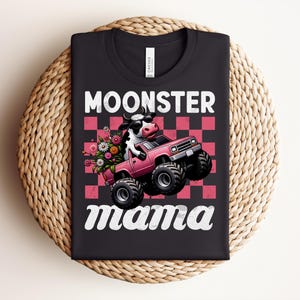 May include: Black t-shirt with the words "MOONSTER mama" in white script. The graphic features a cow driving a pink monster truck with flowers in the back. The background is a pink and red checkerboard pattern.