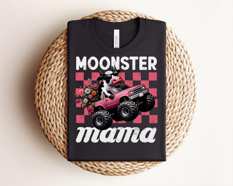 May include: Black t-shirt with the words "MOONSTER mama" in white script. The graphic features a cow driving a pink monster truck with flowers in the back. The background is a pink and red checkerboard pattern.