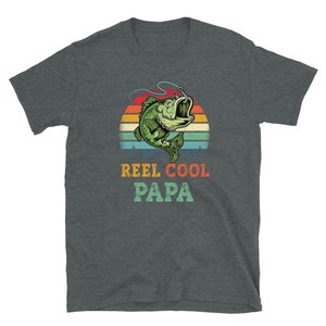 Pu&ograve; includere: Una t-shirt grigio scuro melange con una grafica di tramonto retr&ograve; che presenta un pesce verde con un amo in bocca. Il testo "REEL COOL PAPA" &egrave; stampato sotto la grafica.