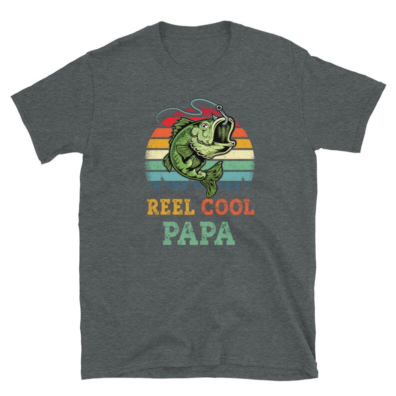 Pu&ograve; includere: Una t-shirt grigio scuro melange con una grafica di tramonto retr&ograve; che presenta un pesce verde con un amo in bocca. Il testo "REEL COOL PAPA" &egrave; stampato sotto la grafica.
