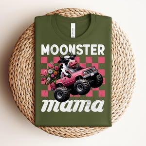 May include: Olive green t-shirt with the words "MOONSTER mama" in white script. The graphic features a cow driving a pink monster truck with flowers in the back, set against a pink and red checkered background.