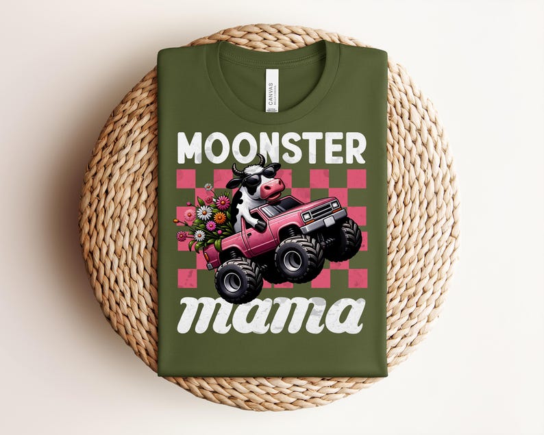 May include: Olive green t-shirt with the words "MOONSTER mama" in white script. The graphic features a cow driving a pink monster truck with flowers in the back, set against a pink and red checkered background.