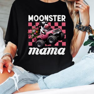 May include: Black t-shirt with the text "MOONSTER mama" in white script. The graphic features a cow driving a pink monster truck with flowers in the bed, set against a pink and white checkered background.