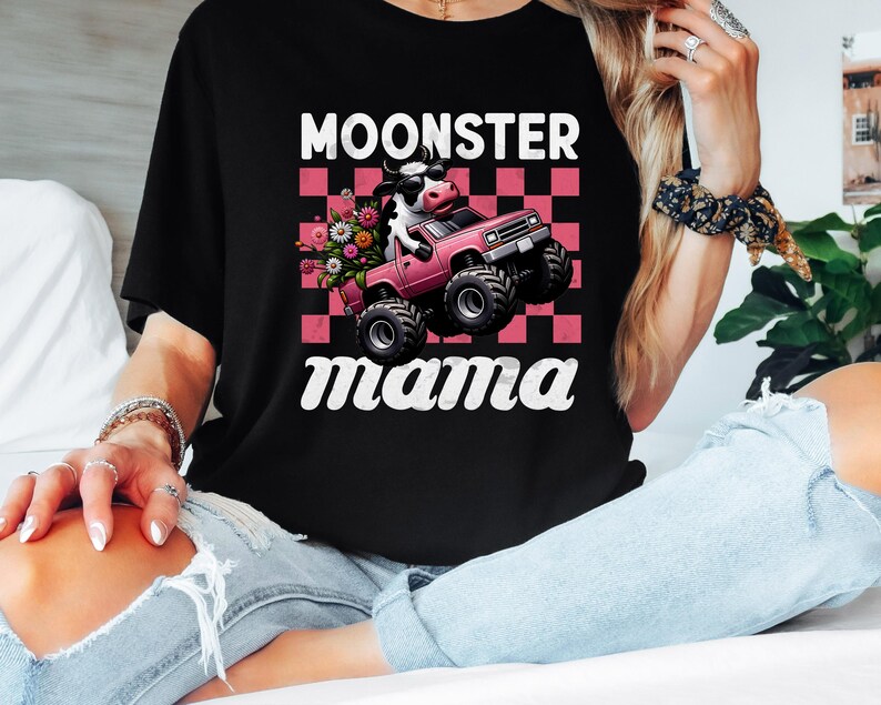 May include: Black t-shirt with the text "MOONSTER mama" in white script. The graphic features a cow driving a pink monster truck with flowers in the bed, set against a pink and white checkered background.