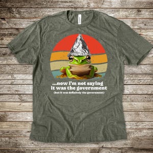 May include: Green t-shirt with a cartoon frog wearing a foil hat, set against a retro sunset design. The text reads, "...now I'm not saying it was the government (but it was definitely the government)."