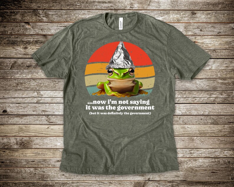 May include: Green t-shirt with a cartoon frog wearing a foil hat, set against a retro sunset design. The text reads, "...now I'm not saying it was the government (but it was definitely the government)."