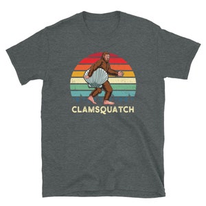 May include: A dark gray t-shirt with a retro sunset graphic featuring a Bigfoot carrying a large seashell. The text "CLAMSQUATCH" is printed below the graphic.