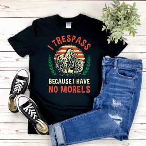 Pu&ograve; includere: T-shirt nero con la scritta "I TRESPASS BECAUSE I HAVE NO MORELS" in rosso e bianco. Il design include una grafica di funghi e uno sfondo ispirato alla bandiera americana. La maglietta &egrave; abbinata a jeans blu e scarpe da ginnastica nere.