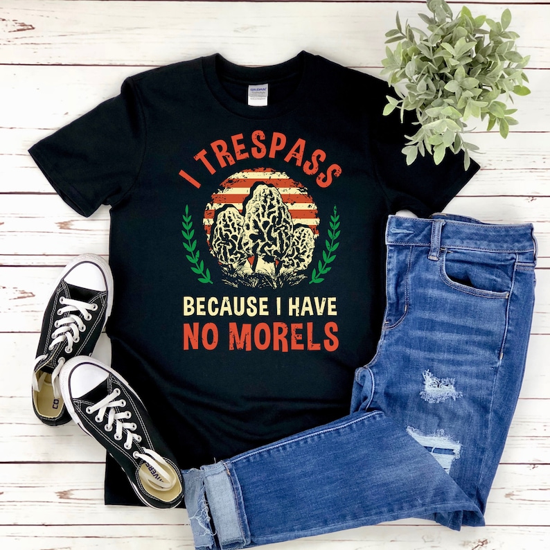 Pu&ograve; includere: T-shirt nero con la scritta "I TRESPASS BECAUSE I HAVE NO MORELS" in rosso e bianco. Il design include una grafica di funghi e uno sfondo ispirato alla bandiera americana. La maglietta &egrave; abbinata a jeans blu e scarpe da ginnastica nere.