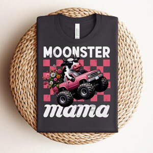 May include: Black t-shirt with the words "MOONSTER mama" in white script. The graphic features a cow driving a pink monster truck with flowers in the back. The background is a pink and red checkerboard pattern.