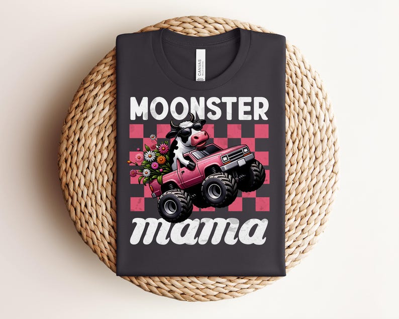 May include: Black t-shirt with the words "MOONSTER mama" in white script. The graphic features a cow driving a pink monster truck with flowers in the back. The background is a pink and red checkerboard pattern.