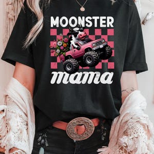 May include: Black t-shirt with the text "MOONSTER mama" in white script. The graphic features a cow wearing sunglasses driving a pink monster truck with a bouquet of flowers in the bed of the truck, set against a pink and red checkered background.