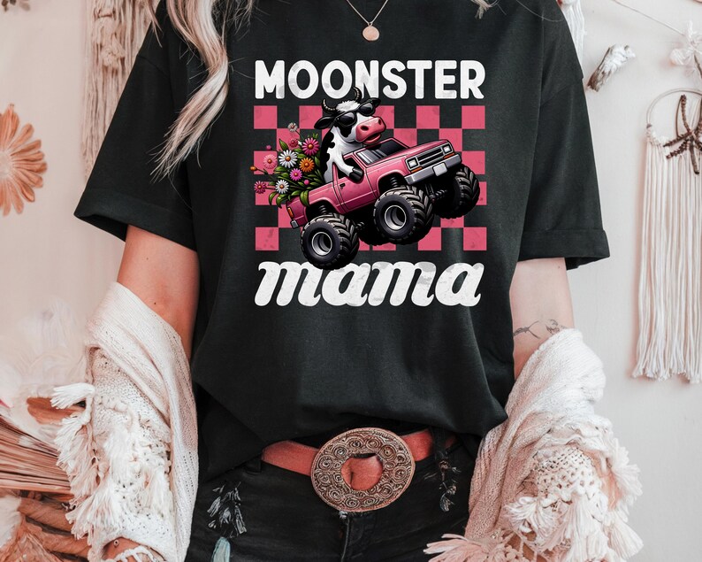 May include: Black t-shirt with the text "MOONSTER mama" in white script. The graphic features a cow wearing sunglasses driving a pink monster truck with a bouquet of flowers in the bed of the truck, set against a pink and red checkered background.