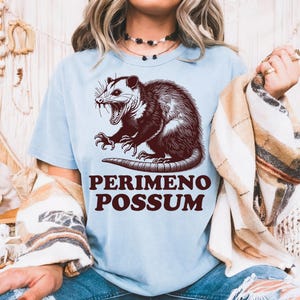 May include: Light blue t-shirt featuring a detailed illustration of an opossum with the text "PERIMENO POSSUM" in a brown, vintage font. The opossum is depicted with its mouth open, showcasing its teeth.