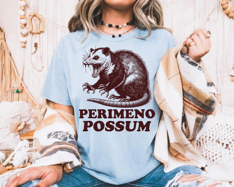 May include: Light blue t-shirt featuring a detailed illustration of an opossum with the text "PERIMENO POSSUM" in a brown, vintage font. The opossum is depicted with its mouth open, showcasing its teeth.