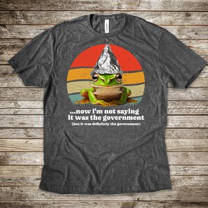 May include: Dark grey t-shirt with a cartoon frog wearing a foil hat, set against a retro sunset design. The shirt includes the text: "...now I'm not saying it was the government (but it was definitely the government)".
