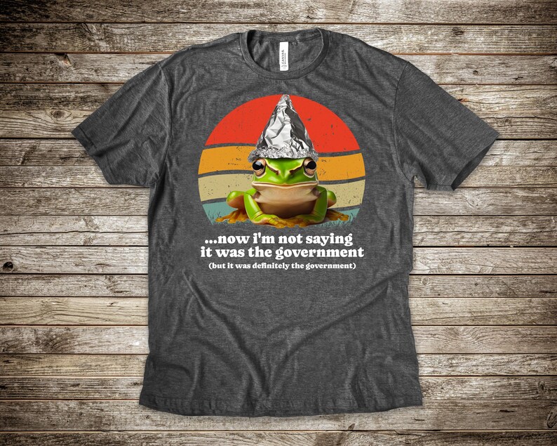 May include: Dark grey t-shirt with a cartoon frog wearing a foil hat, set against a retro sunset design. The shirt includes the text: "...now I'm not saying it was the government (but it was definitely the government)".