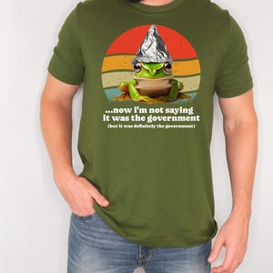 May include: Olive green t-shirt with a cartoon frog wearing a foil hat, set against a retro sunset design. The shirt includes the text: "...now I'm not saying it was the government (but it was definitely the government)".